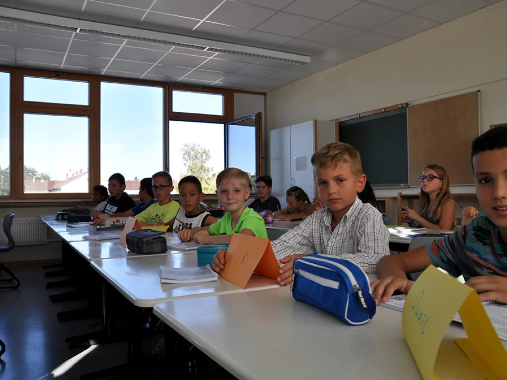 Kinder sitzen an zusammengestellten Tischen und schauen in die Kamera.